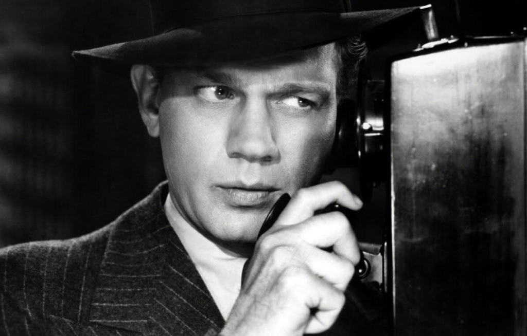 A man presses his ear to a phone in a phone booth. He is wearing a pinstripe blazer and a fedora.